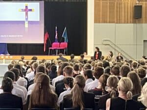 A crowded school auditorium facing a stage with a large screen showing a cross and three pink chairs on stage behind flags.