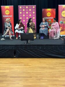 Panel of five women seated on a stage with colorful festival banners behind them, participating in a discussion.